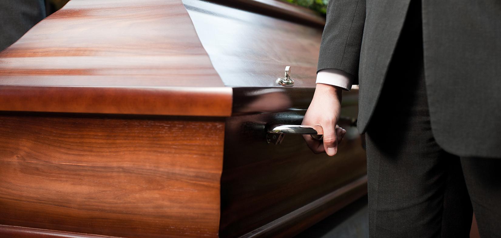 Nahaufnahme eines Anzugs tragenden Mannes, der den Griff eines hölzernen Sargs hält, mit Blumen auf dem Sargdeckel.