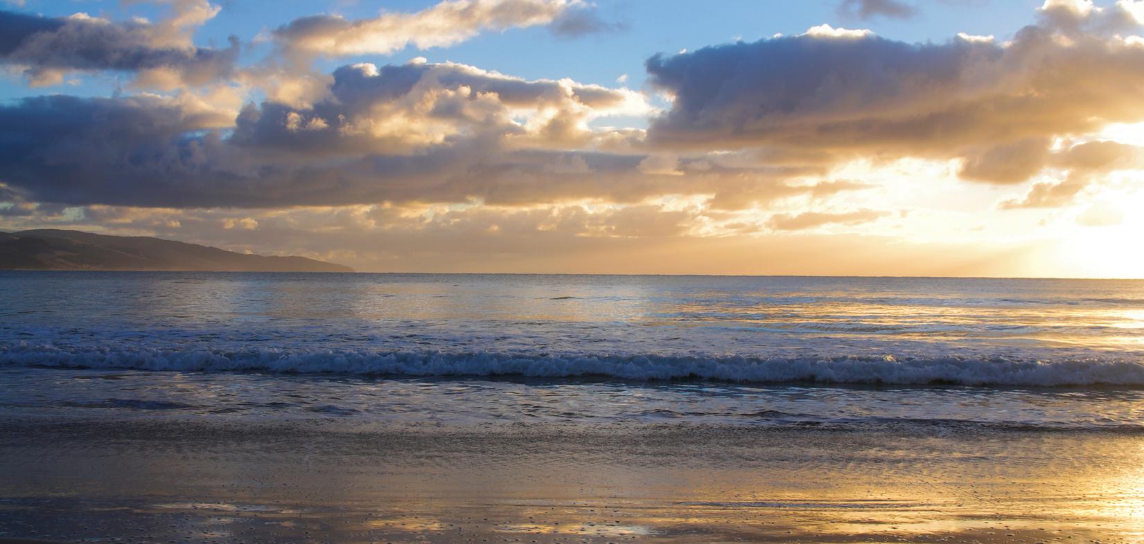 Sonnenuntergang über dem Meer, sanfte Wellen brechen am ruhigen Strand, Wolken reflektieren das Licht am Himmel.