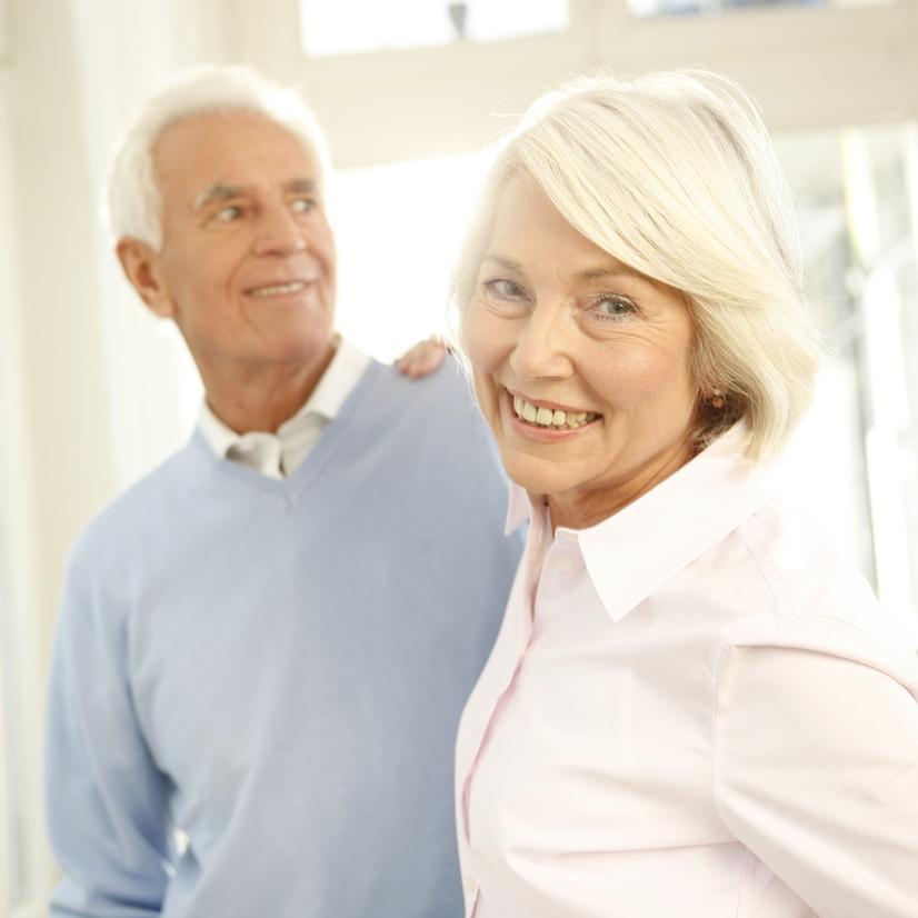 Ein lächelndes älteres Paar steht vor einem Fenster, umgeben von hellem Licht, mit sanften Farben im Hintergrund.
